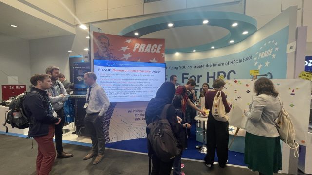 A front view of the PRACE booth with a circular canopy above, a digital screen on the left and our comunity artwork on the right. The digital screen shows a presentation about PRACE. The community artwork consists of colourful stickers in various shapes on a white canvas. People are standing inside and outside the booth, talking and placing stickers on the canvas.